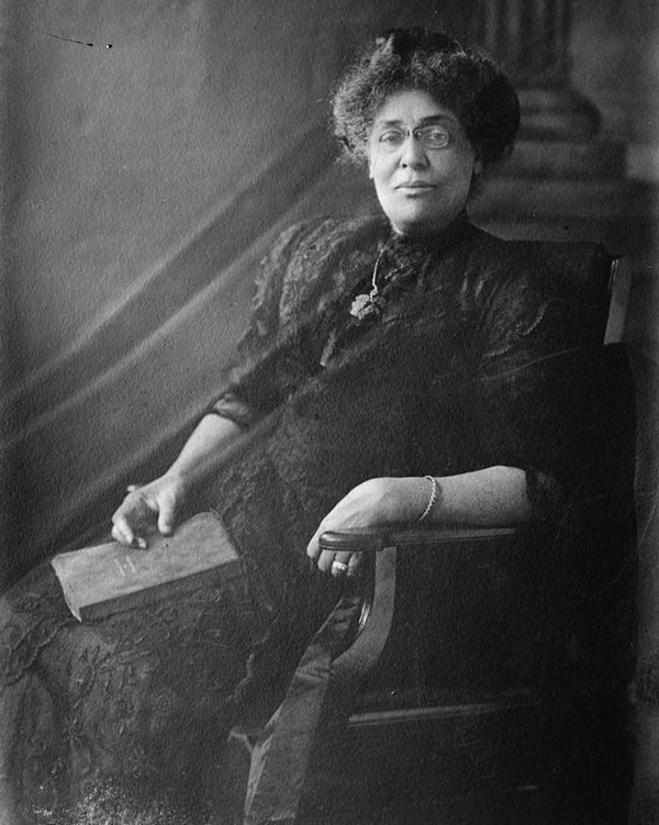 Black woman sitting at a chair with a book on her lap. She wears a black lacy dress and glasses.
