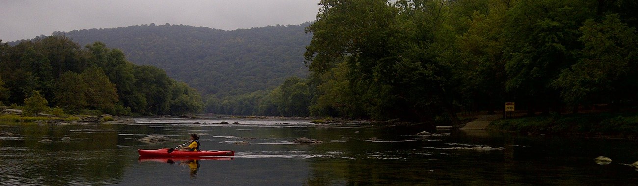kayaker on a calm, wide river with forested hillsides on both shorelines