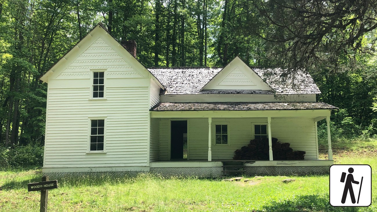 A white historic home in a grassy field with forest behind it. A wooden sign labels the home.