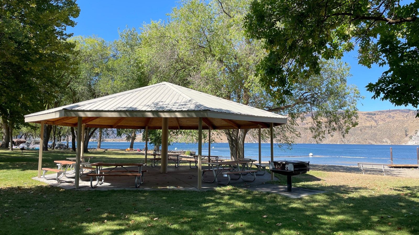 A pointed pergola covers picnic benches on a grassy area near a beach and swim area.