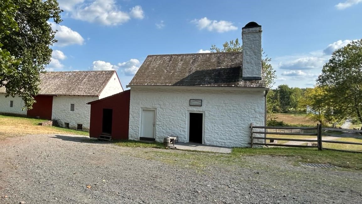 A white and red building with a door open set slightly off a gravel path between larger buildings. 