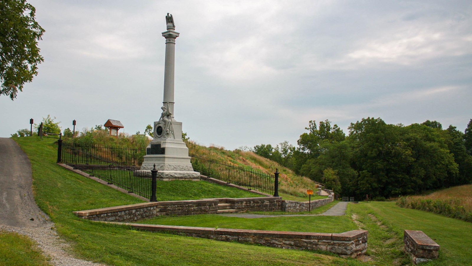 A tall white marble monument sits halfway up a hilltop adjacent to a gravel drive.