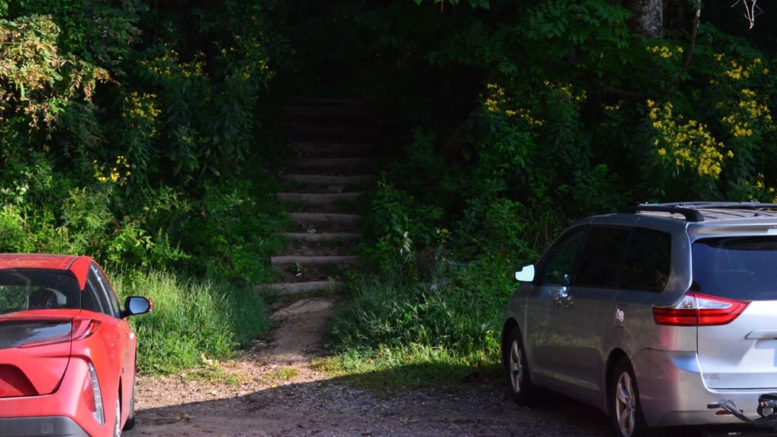 Cars parked in gravel spaces in front of trailhead steps heading into woods