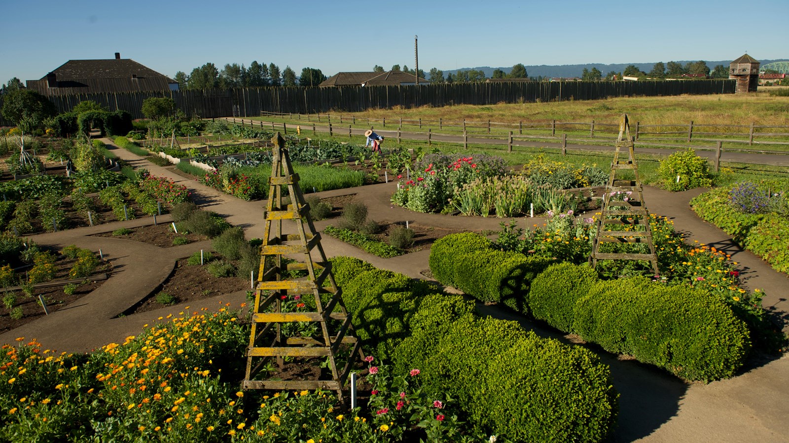 A lush garden with paved pathways. Fort Vancouver can be seen in the background.