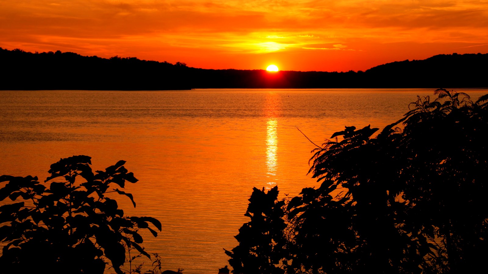 The sun dips behnd a silhouette of rolling hills, turning the lake and sky a brilliant orange.