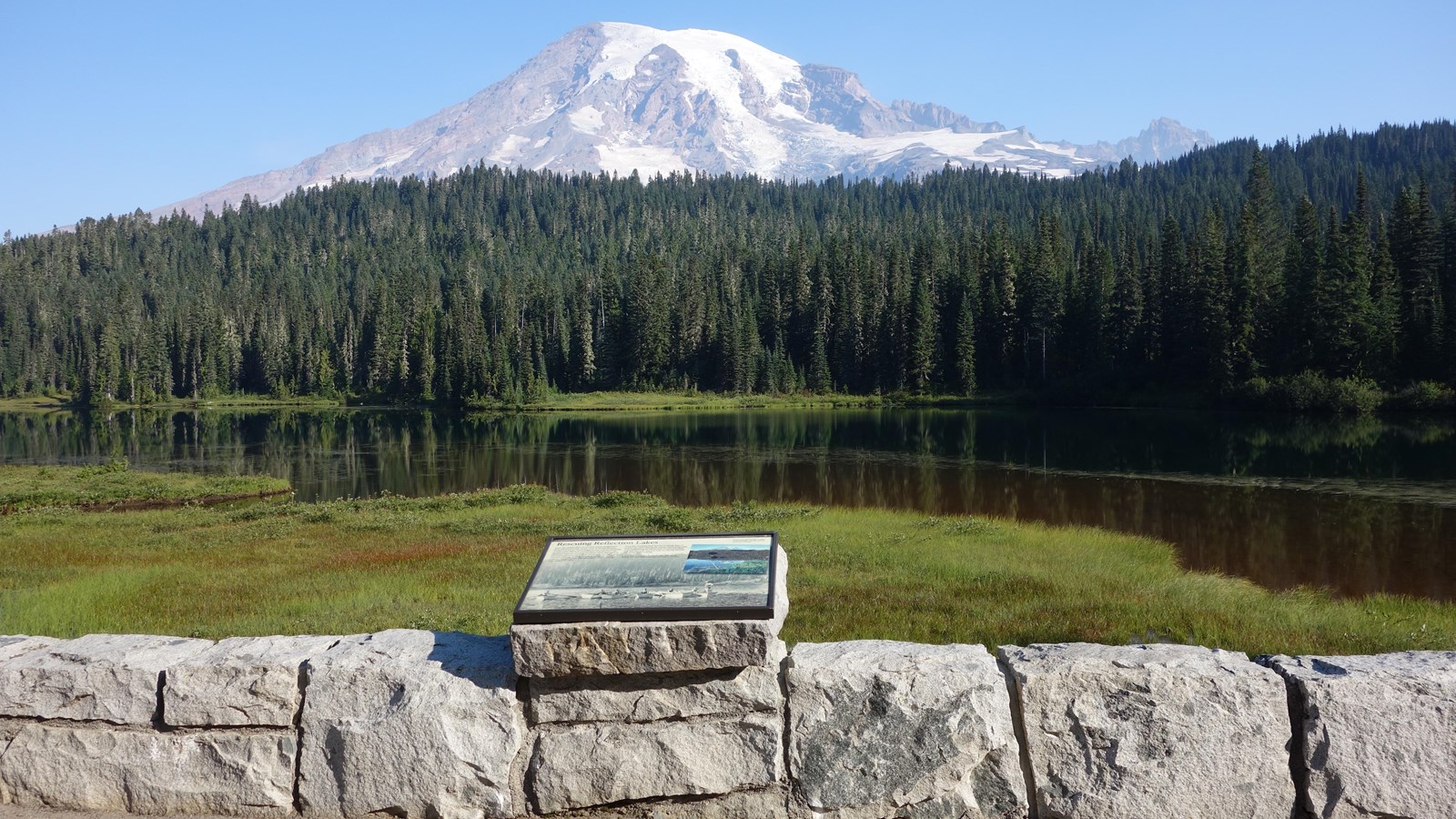 A wayside panel attached to a low rock wall overlooks a lake and view of Mount Rainier.