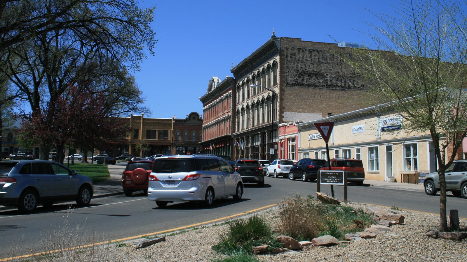 Cars on a city street, next to a center park and historic buildings.
