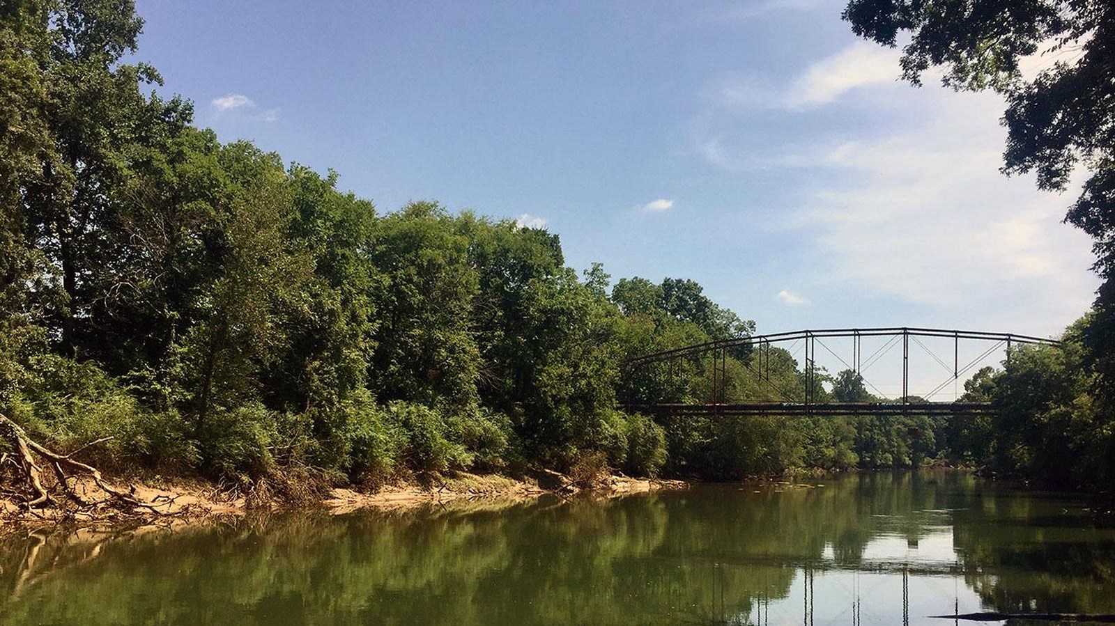 Remains of steel bridge crossing the Chattahoochee River with wooded shorelines and blue sky.