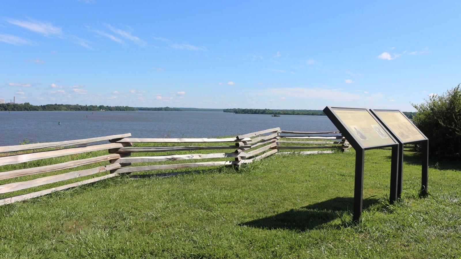 view with blue sky, river beneath, and green grass in the foreground and two signs on the right side