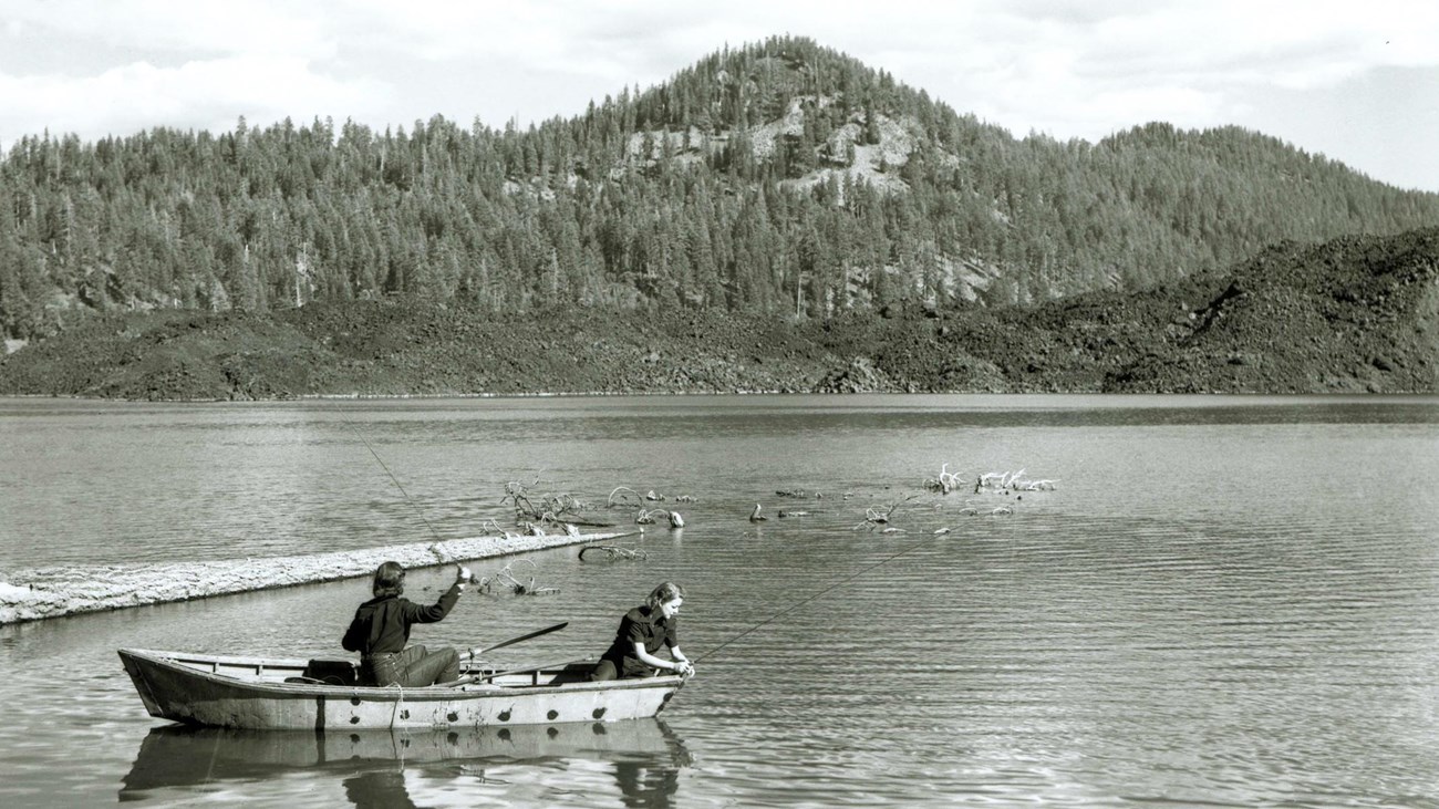 Two women fishing from a row boat on a lake backed by a lava flow and a volcanic peak.