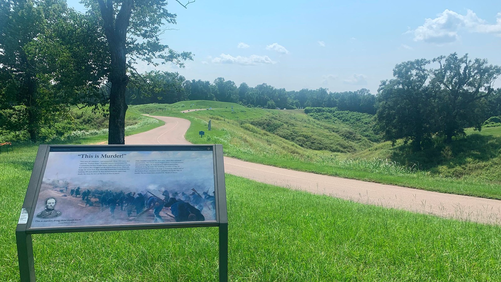 A wayside marker standing on a green grassy area with a road running across the picture.