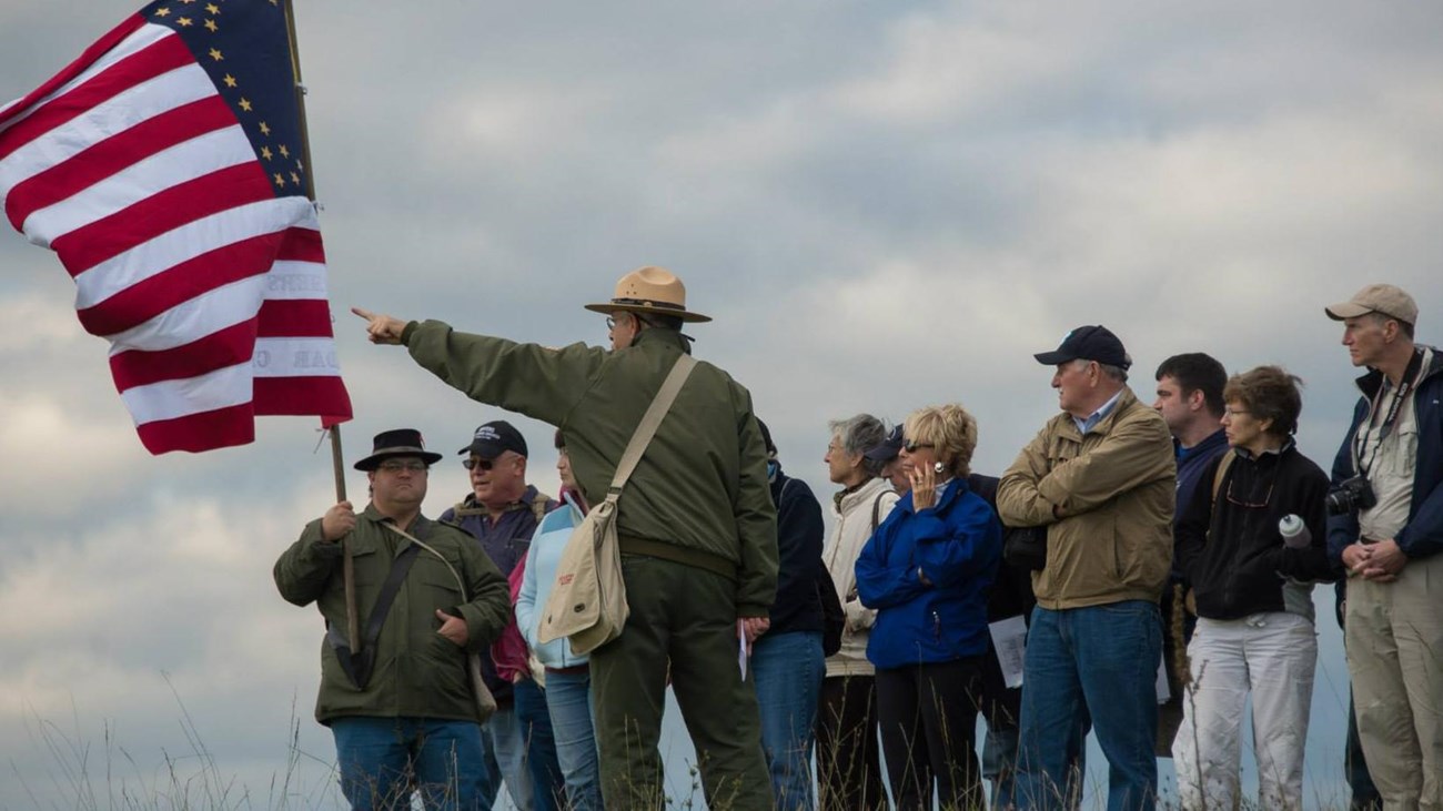 A ranger with visitors on a grass field points past a US flag held by a Civil War reenactor.