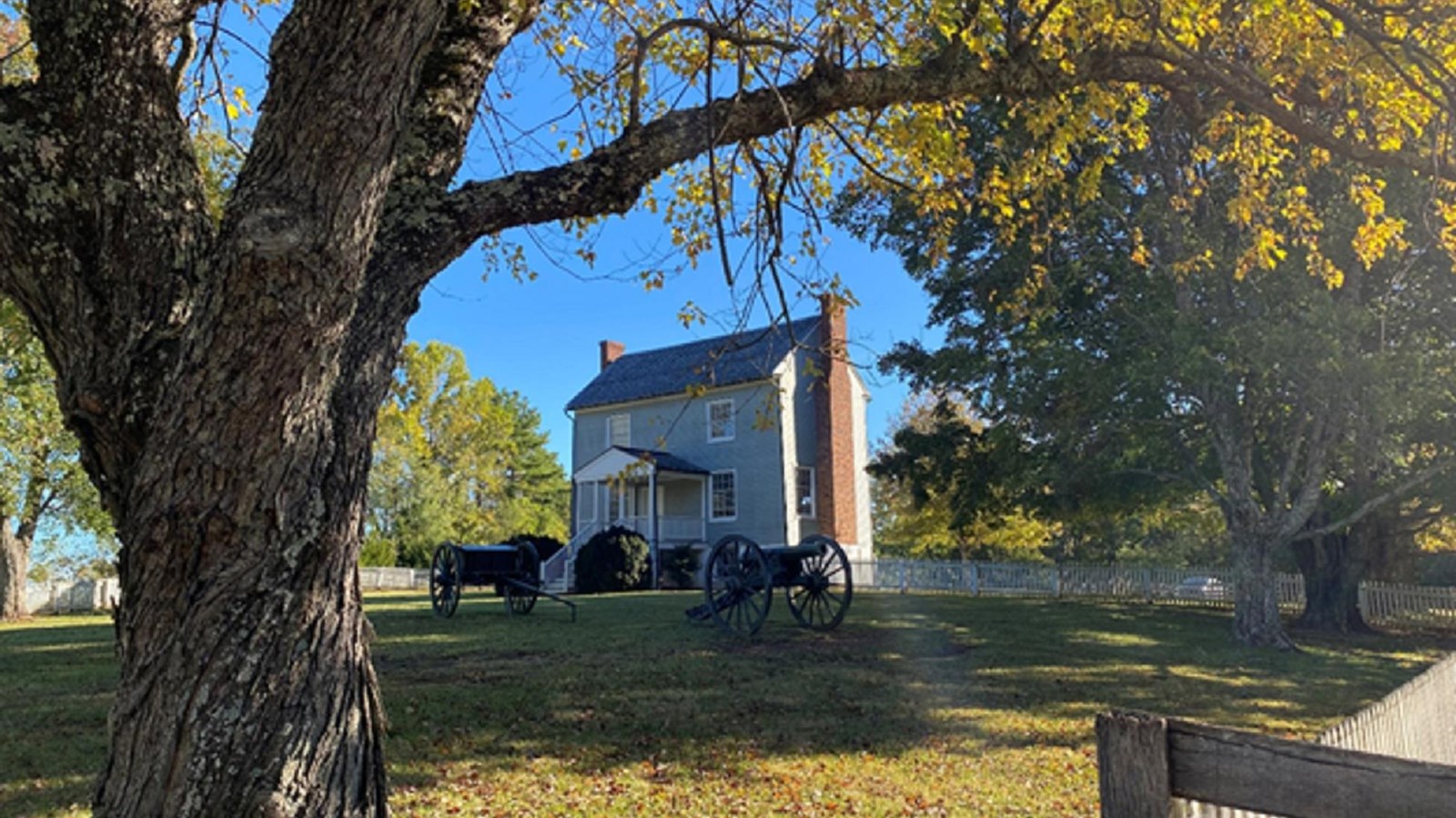 A gray two-story home with large trees and a cannon and limber in the foreground. 