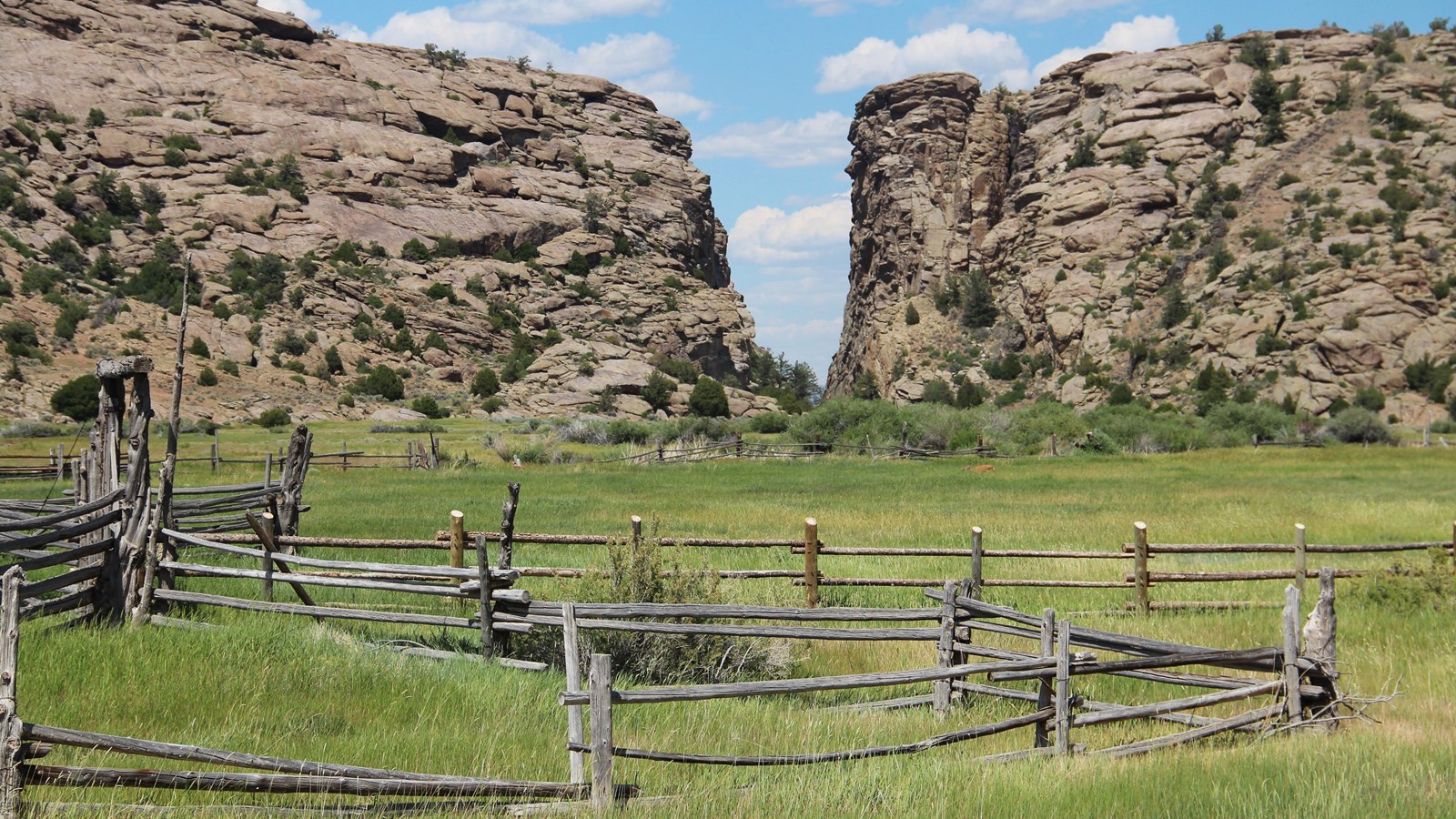 Corral on grassy valley; cracked brown granite cliffs split by V-notch, blue sky with clouds. 