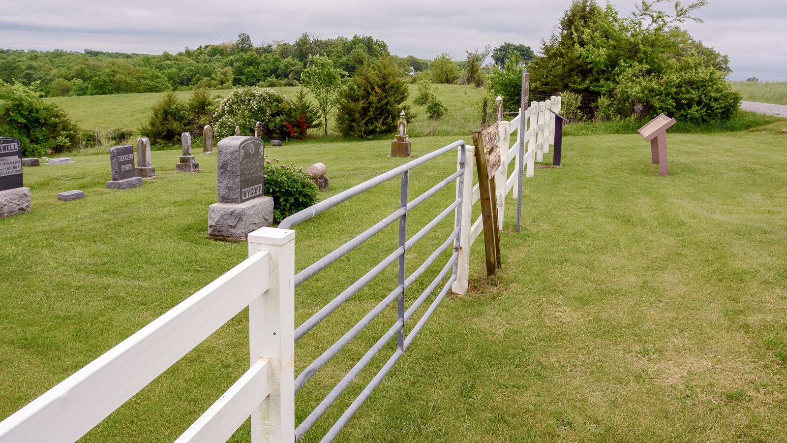 White fence in middle of photo, grass to the right side and a cemetery with headstones to the left.