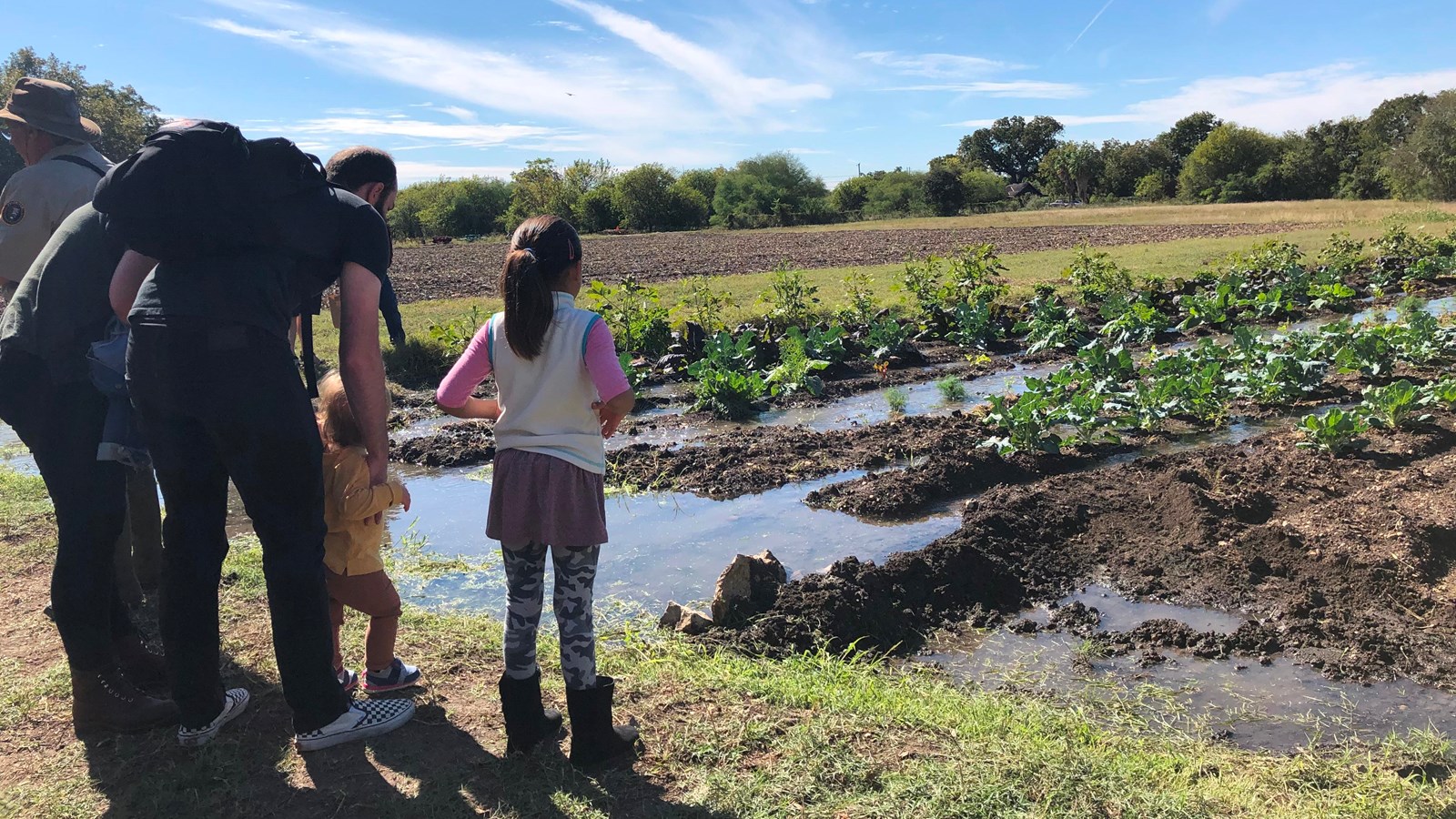 Girl stands with her family looking out toward rows of agriculture being irrigated by an acequia.