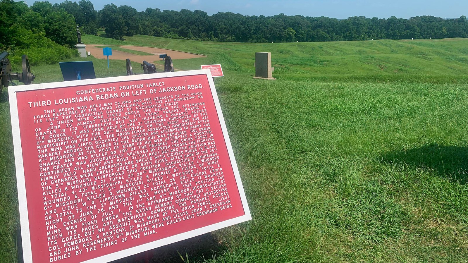 A red iron square tablet stands on a green grassy hill with two cannon and monuments in the back.