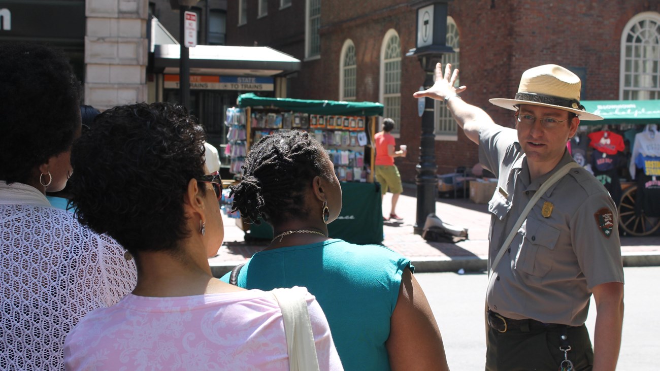 A White park ranger hand raised towards a historic building speaking to a diverse tour group.