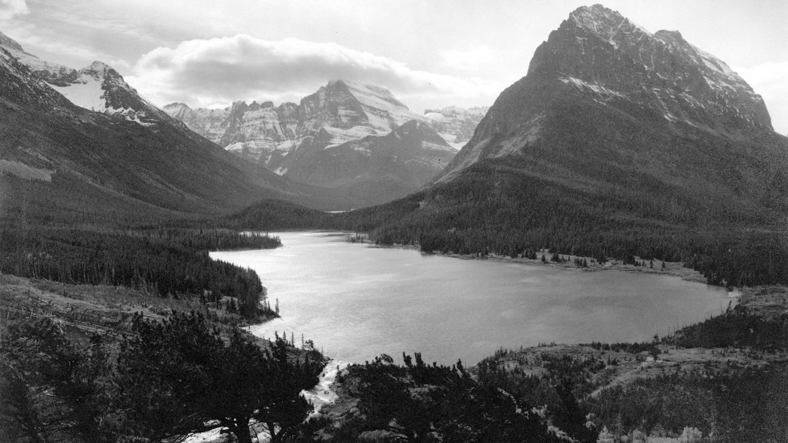 Historic photograph of snowy mountain range surrounding a lake that empties via waterfall. No roads 