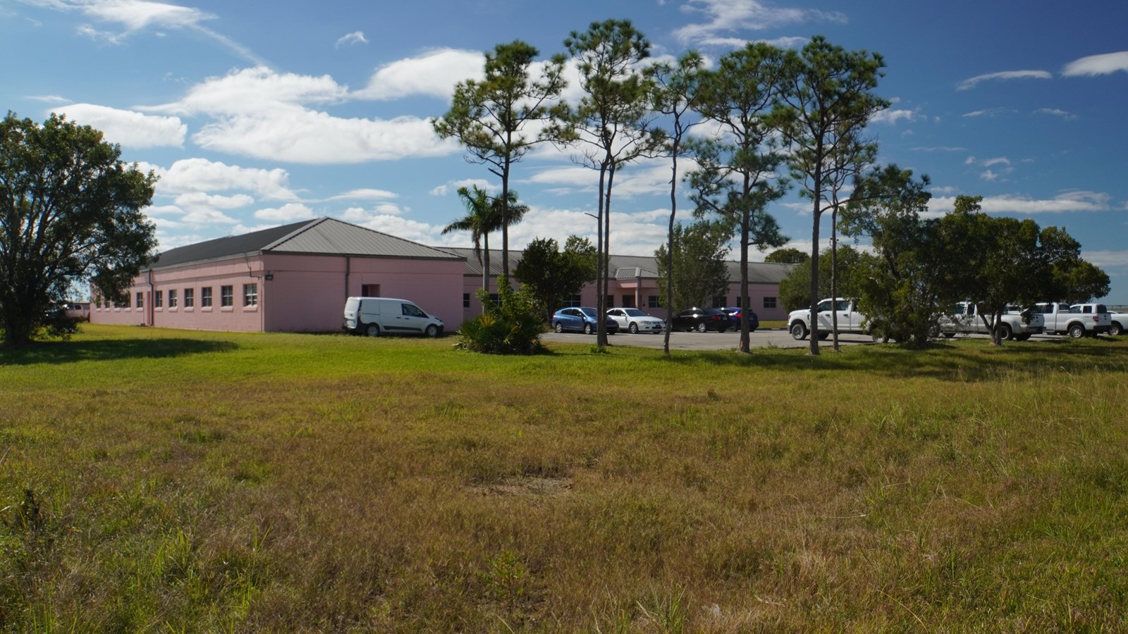 A single storied pink building with a gray roof. Pine trees surround a parking lot with vehicles