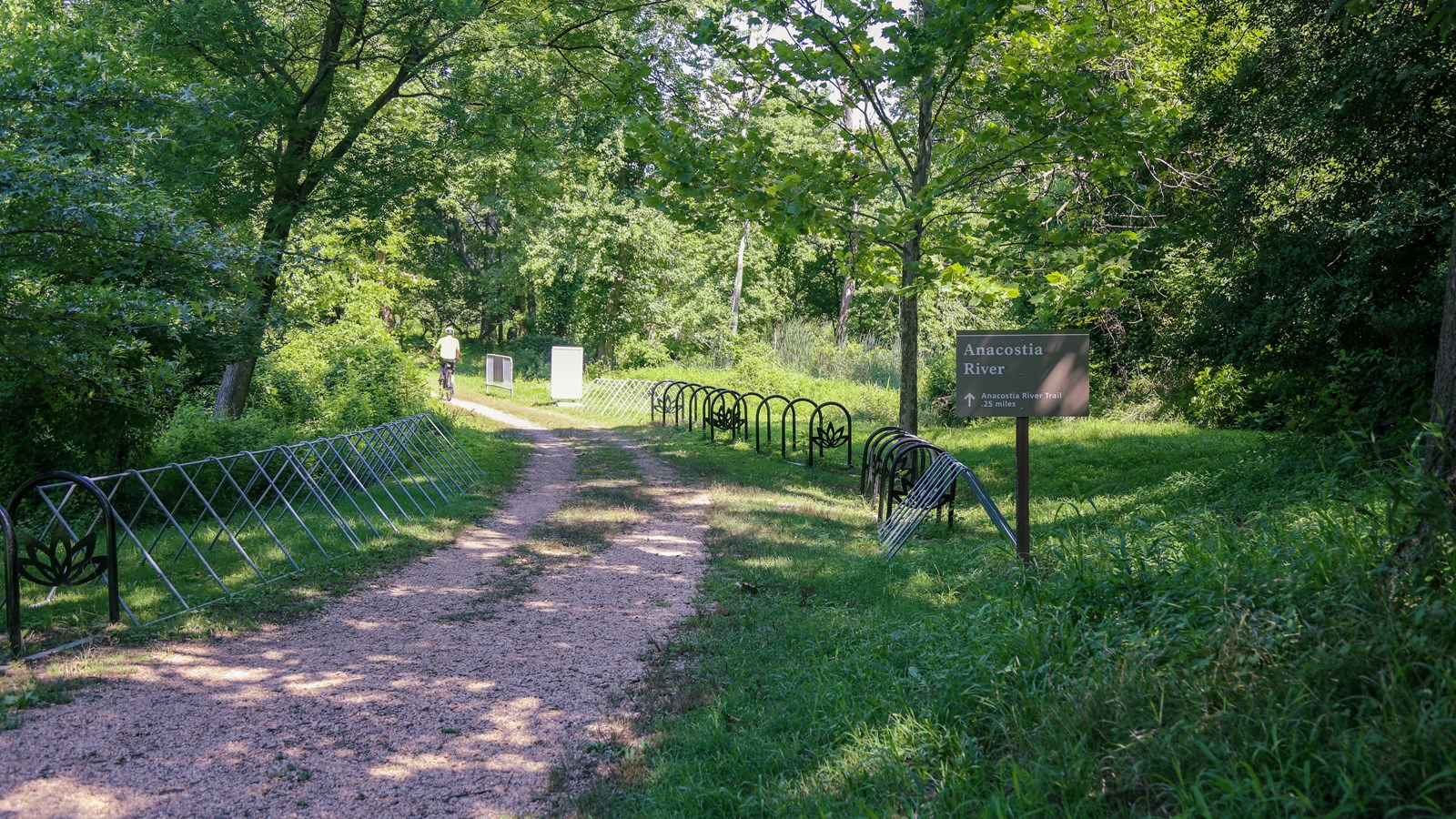 A dirt trail is positioned in between two sets of bike rakes and a trailhead sign. 
