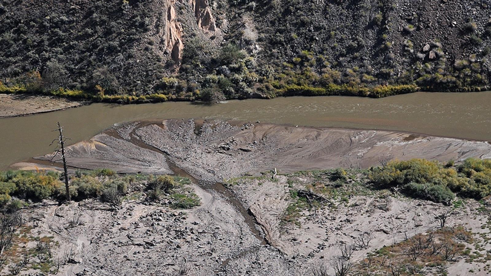 A creek winds along a muddy plain and meets a wider brown river with cliffs in the background.