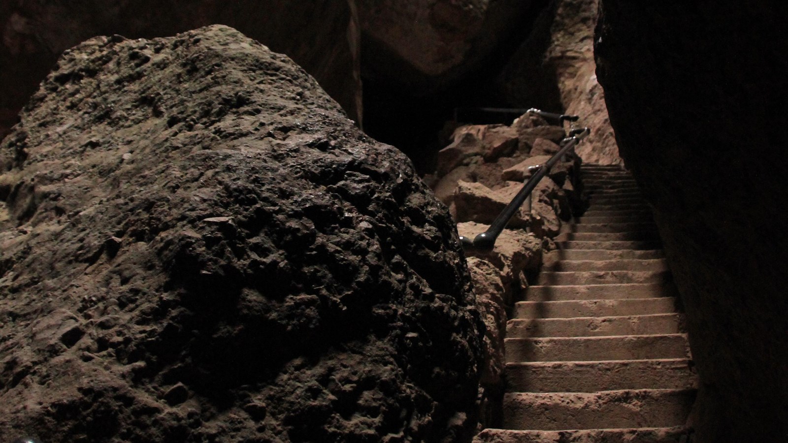 View of a large boulder and rock carved steps adjacent a handrail underneath a rock roofed canyon.