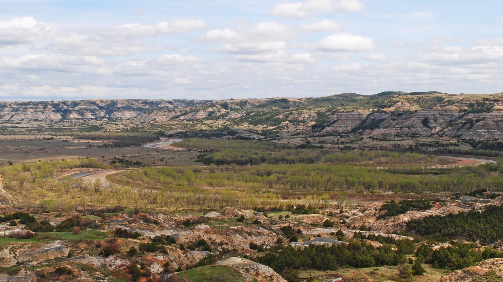 A river meanders through cottonwood trees and steep buttes. 