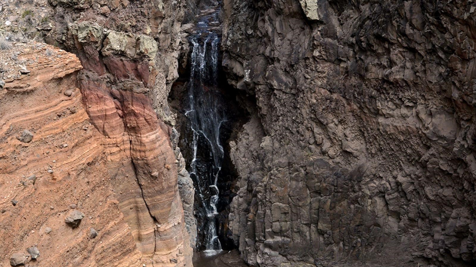 A waterfall pours down dark rock, with lighter tan rock to one side.