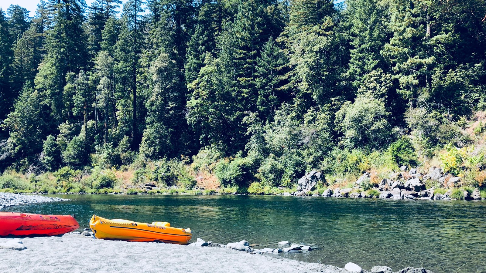 Two adults on a red inflatable kayak. Redwood forests cover the hills either side of a green river.