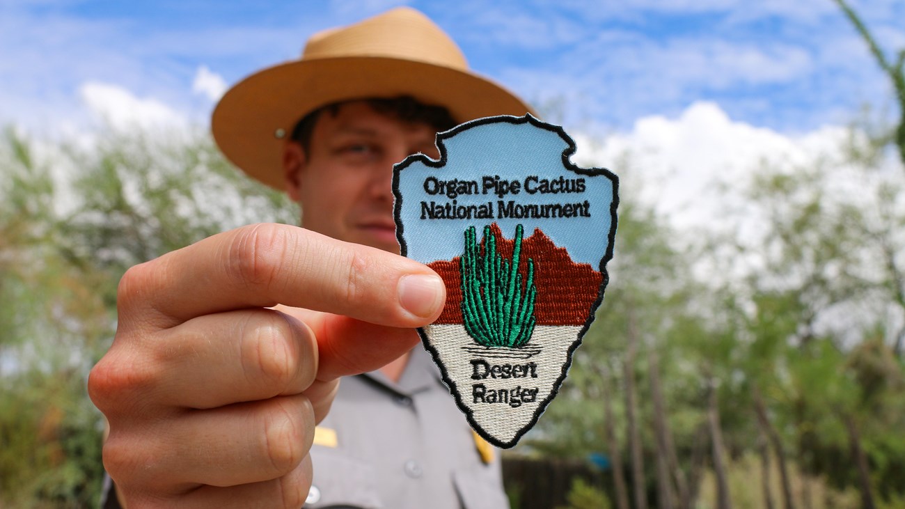 Ranger holding a Desert Ranger patch up to the camera. Native cacti are in the background.