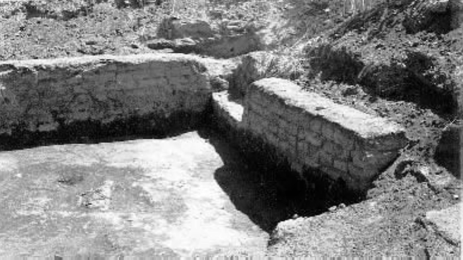 A black and white photo of the corner where two short adobe brick walls meet surrounded by dirt