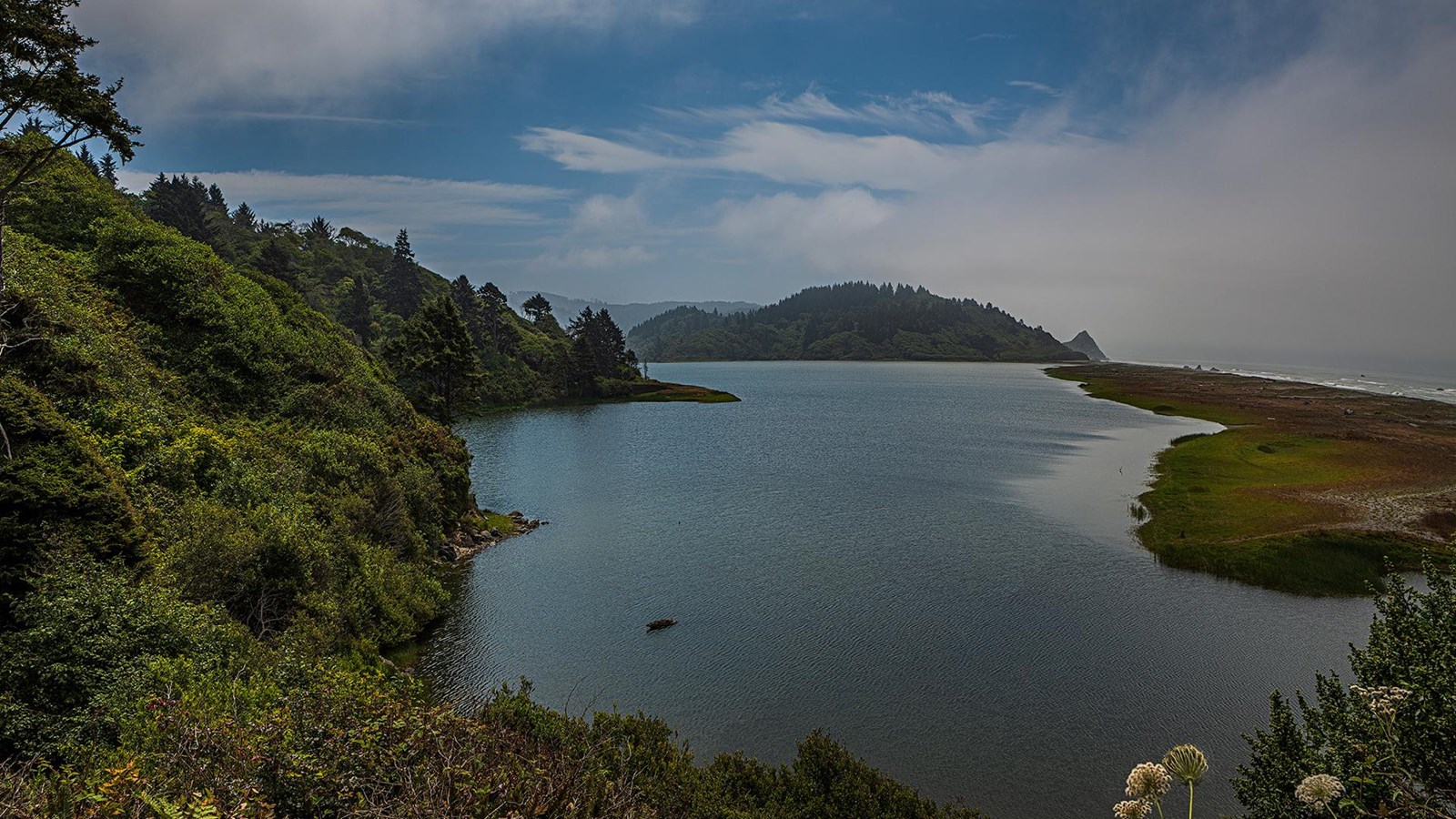 A calm lagoon with blue water. Hills with trees on the left, a spit of sand and ocean to the right.