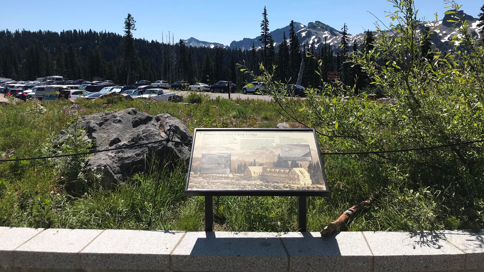 A wayside panel in a metal frame overlooks an island of plants and a parking lot and mountain peaks.