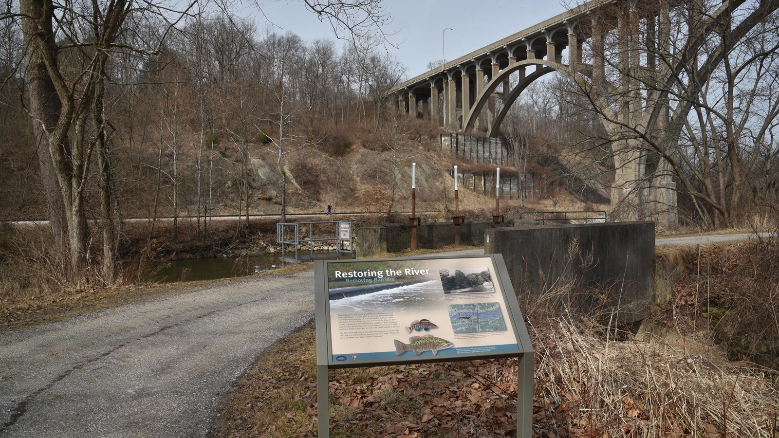 A graphic panel next to a curved paved trail; behind, an arched cement road bridge spans a river.