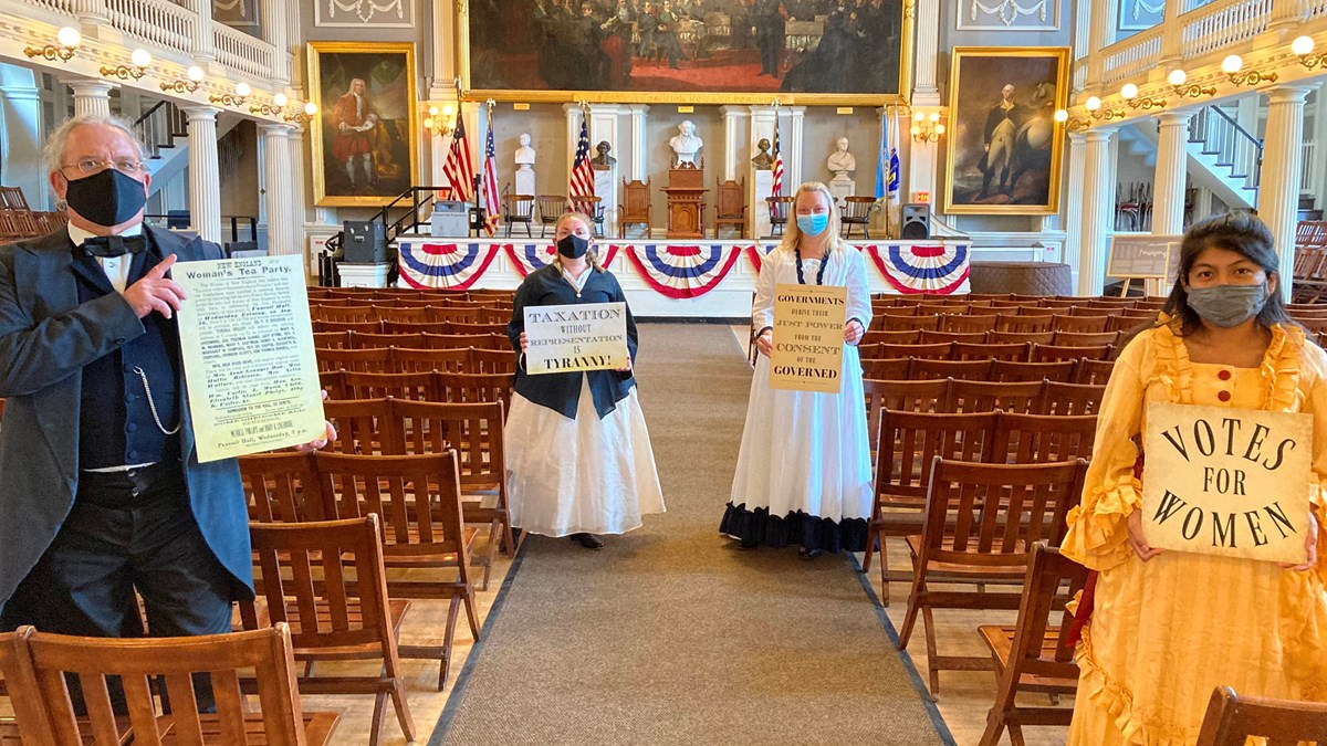 three women and one man in 19th century clothing are standing in the main aisle of Faneuil Hall.
