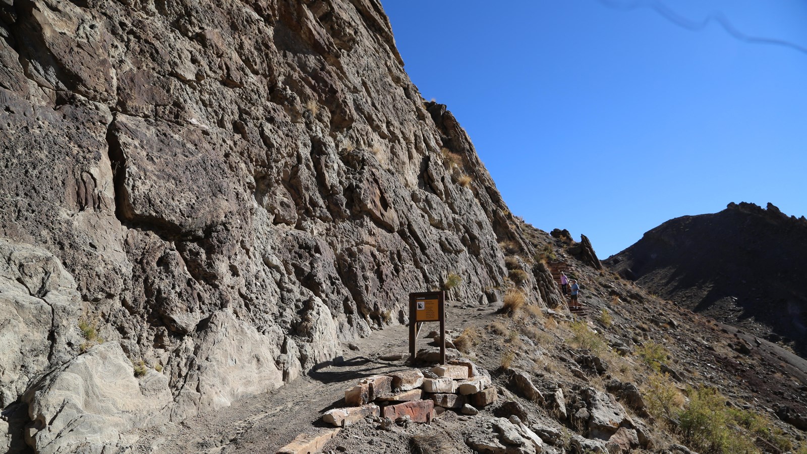 A dark colored sandstone cliff with a trail going alongside.