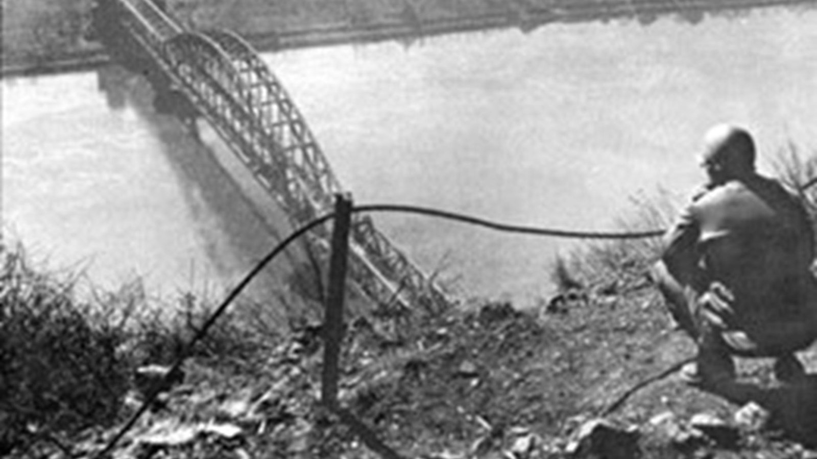 Uniformed man staring down at a large metal structure stretching across a narrow body of water.