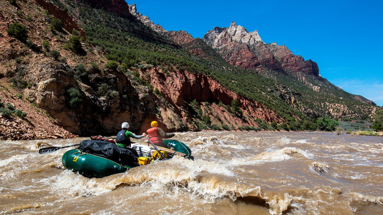 A green raft rides over a whitewater rapid on a brown colored river.