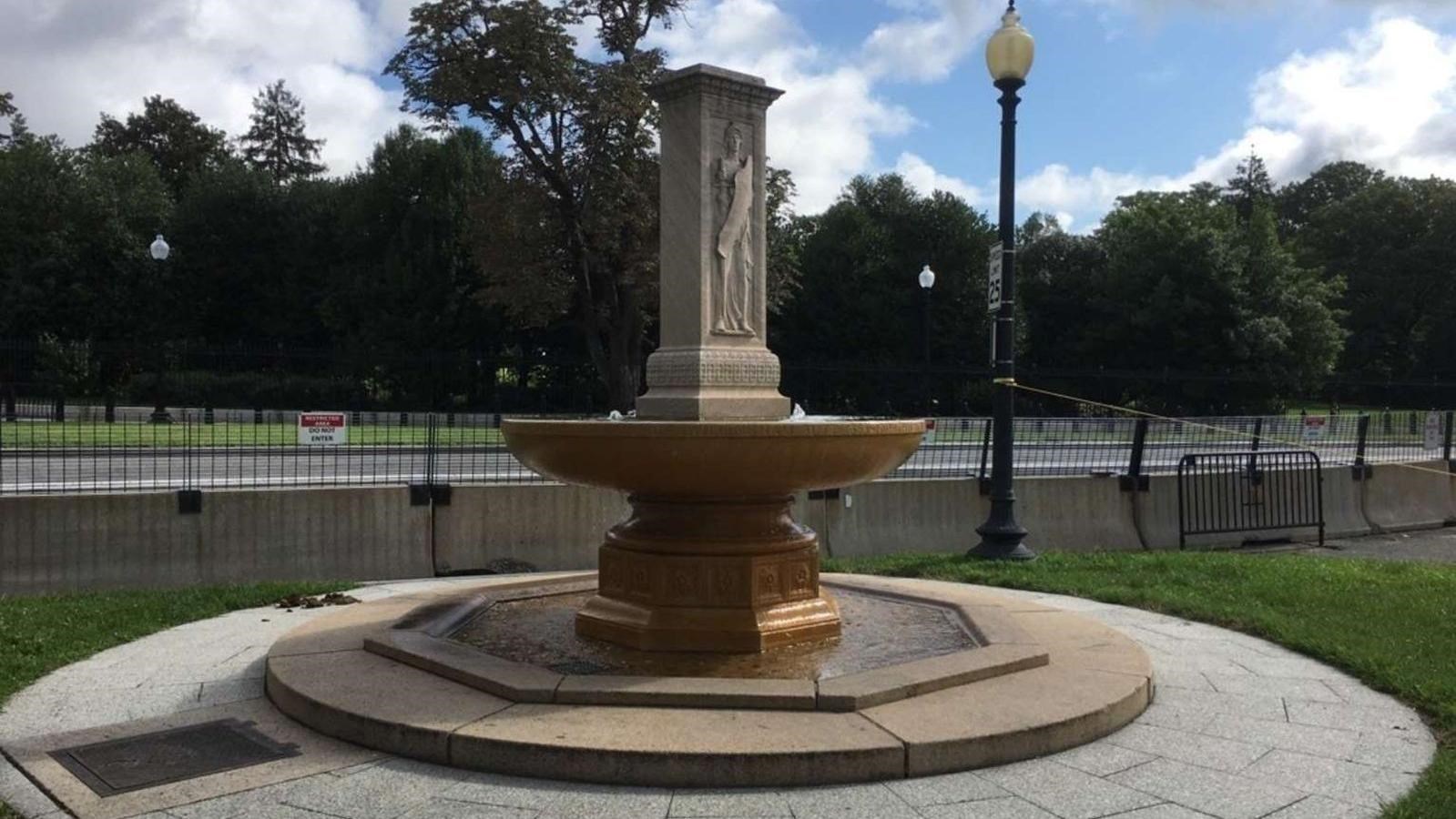 A circular fountain with a rectangular stone sculpture depicting Francis Davis Millet in the middle.