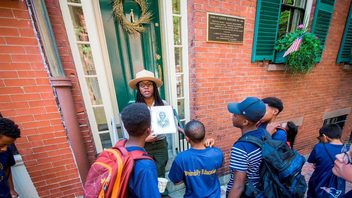 An African American NPS Ranger speaking to a group of children and holding an image of a Black man.