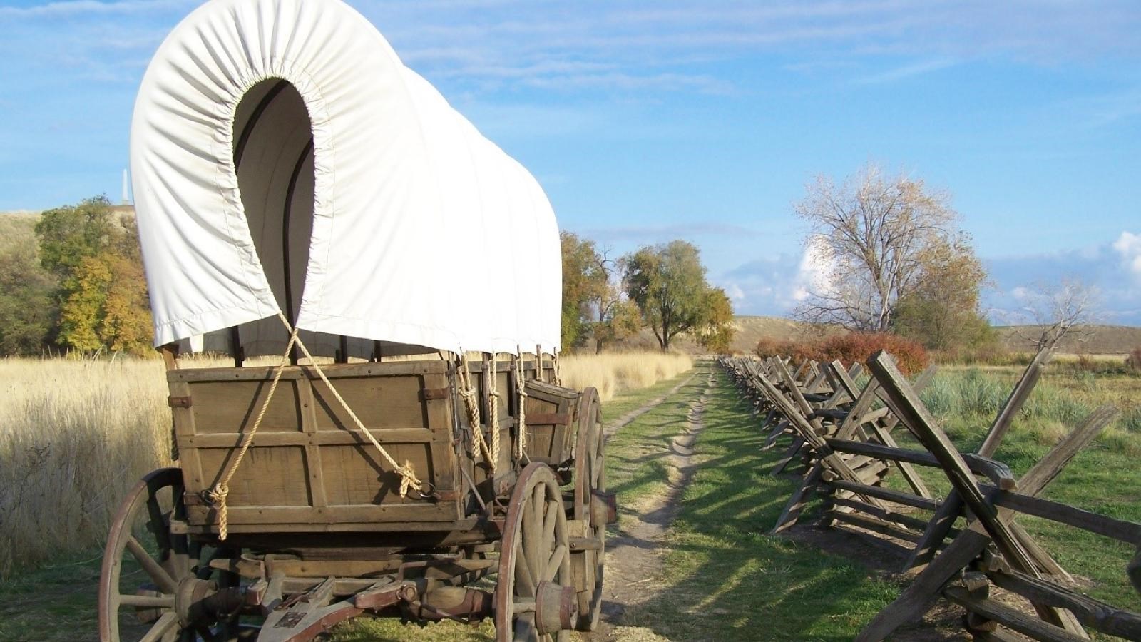 A covered wagon sits on a trail next to a split rail fence and golden tallgrass