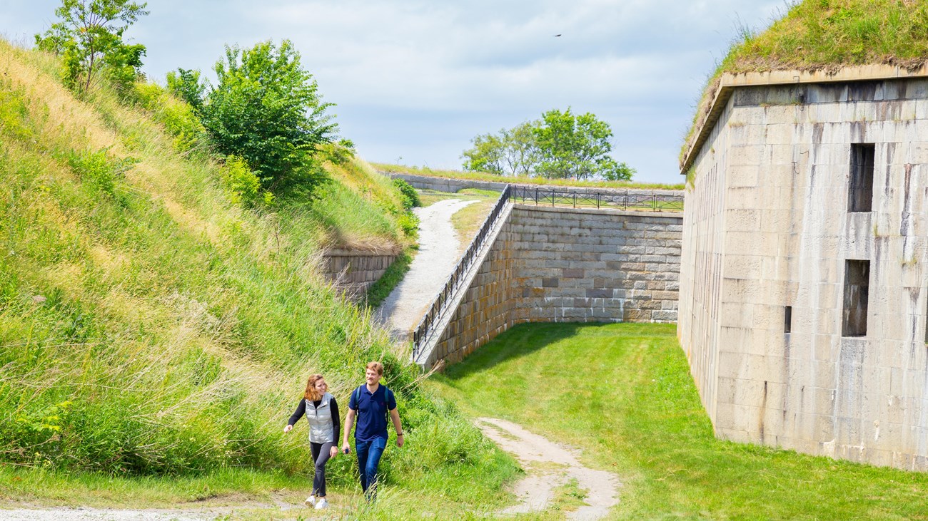 Two people walking on a trail with a fort\'s granite wall to the left of them.