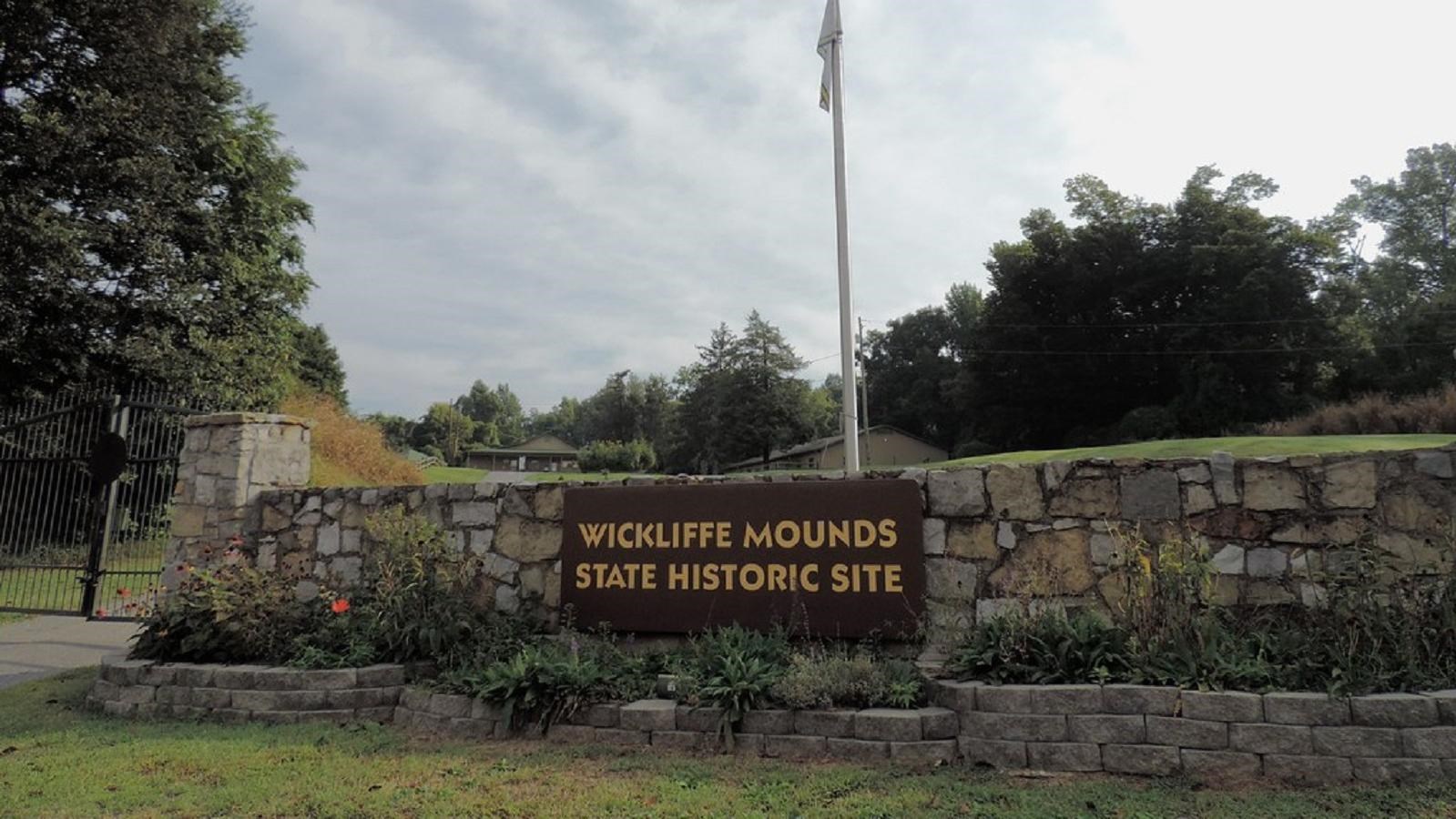 A low stone wall supports a dark sign reading “Wickliffe Mounds State Historic Site”