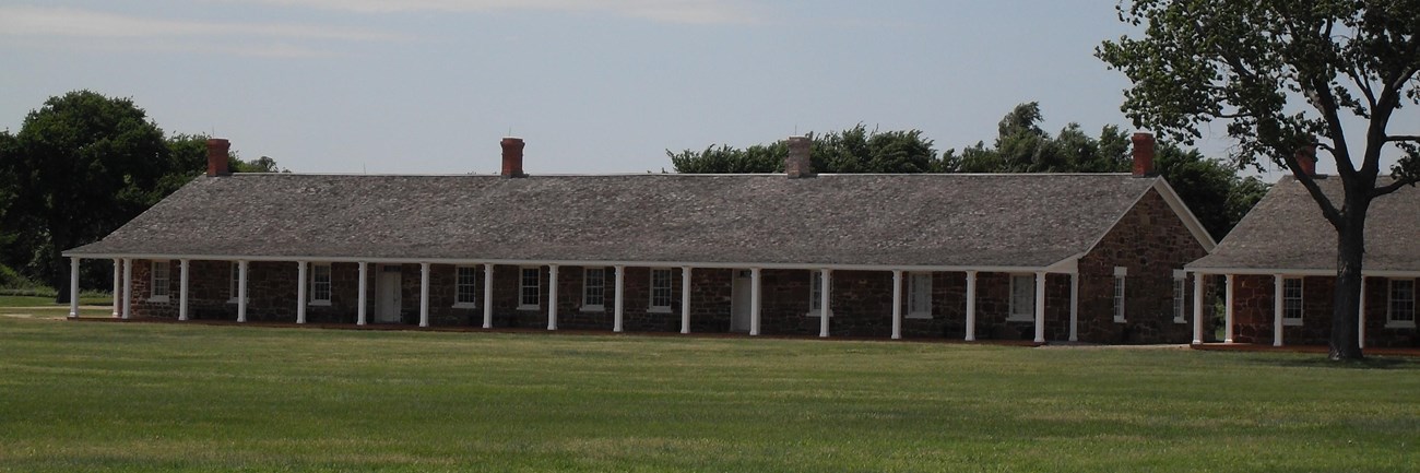 Long distance view of one-story sandstone building with covered porch held up by white columns.