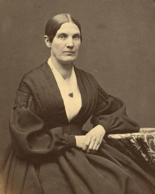 A woman with her hair pulled back wearing a dark dress, seated looking sternly at the camera.