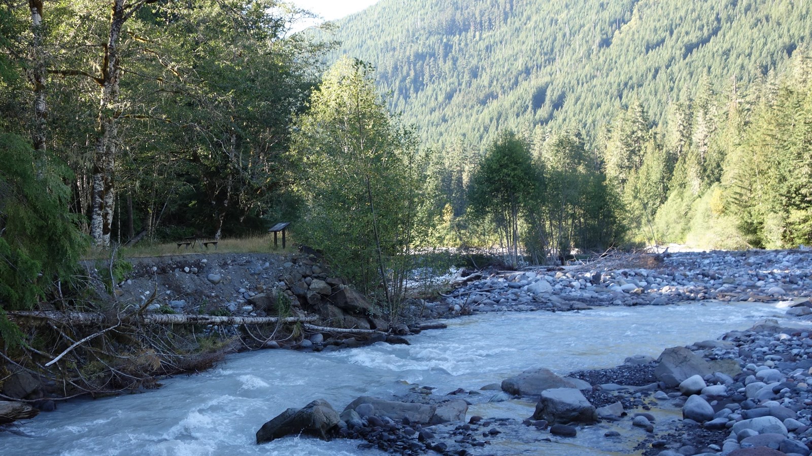 A wayside in a metal frame stands at the edge of a rocky river bed in a forested valley.