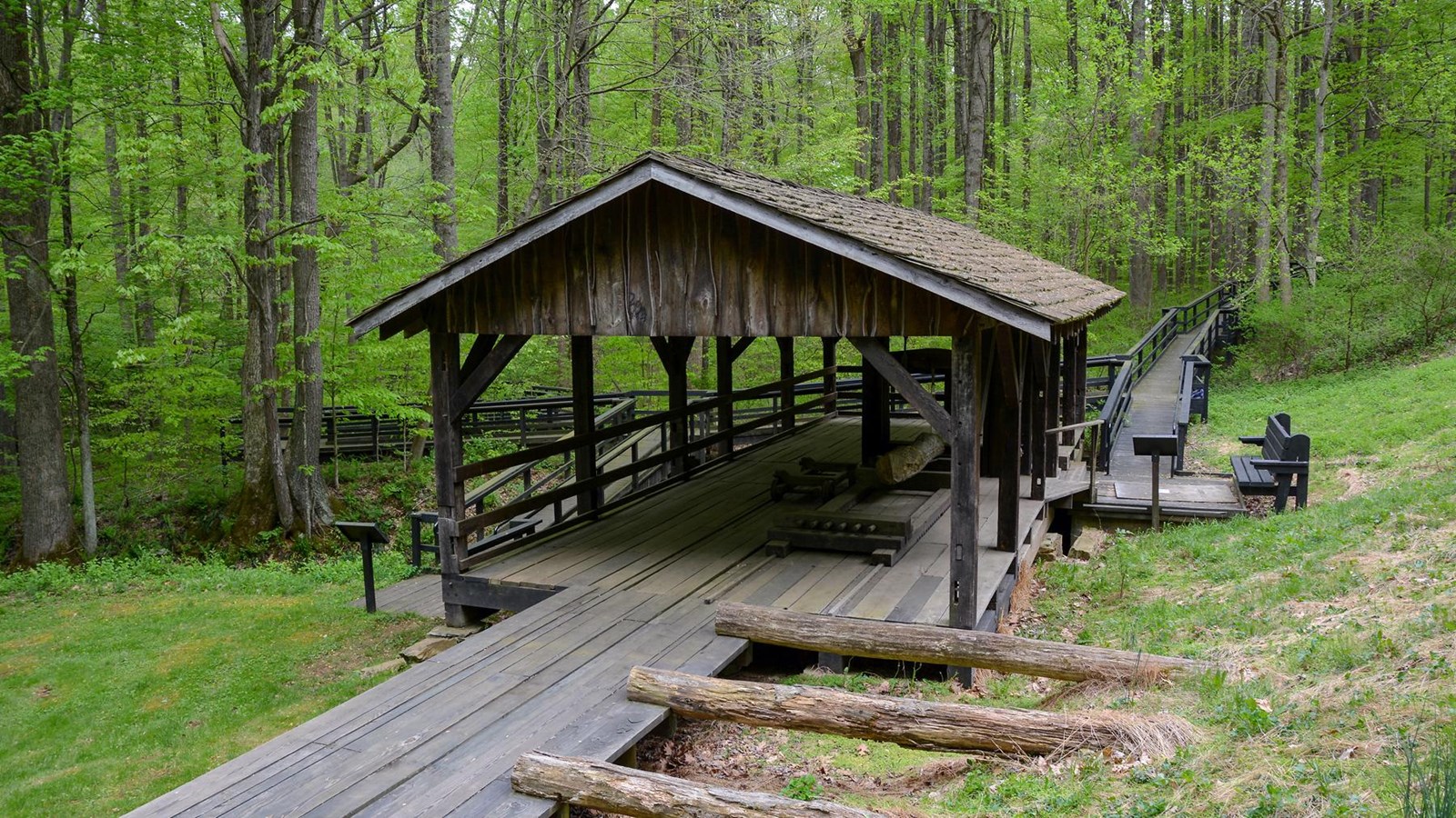 A large open, wooden-roof building with a log and water wheel towards the center. 