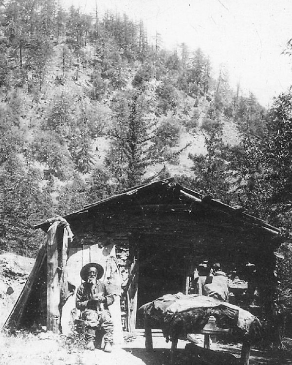 Homesteader sitting in front of a small cabin with various supplies on porch. A mountain background.