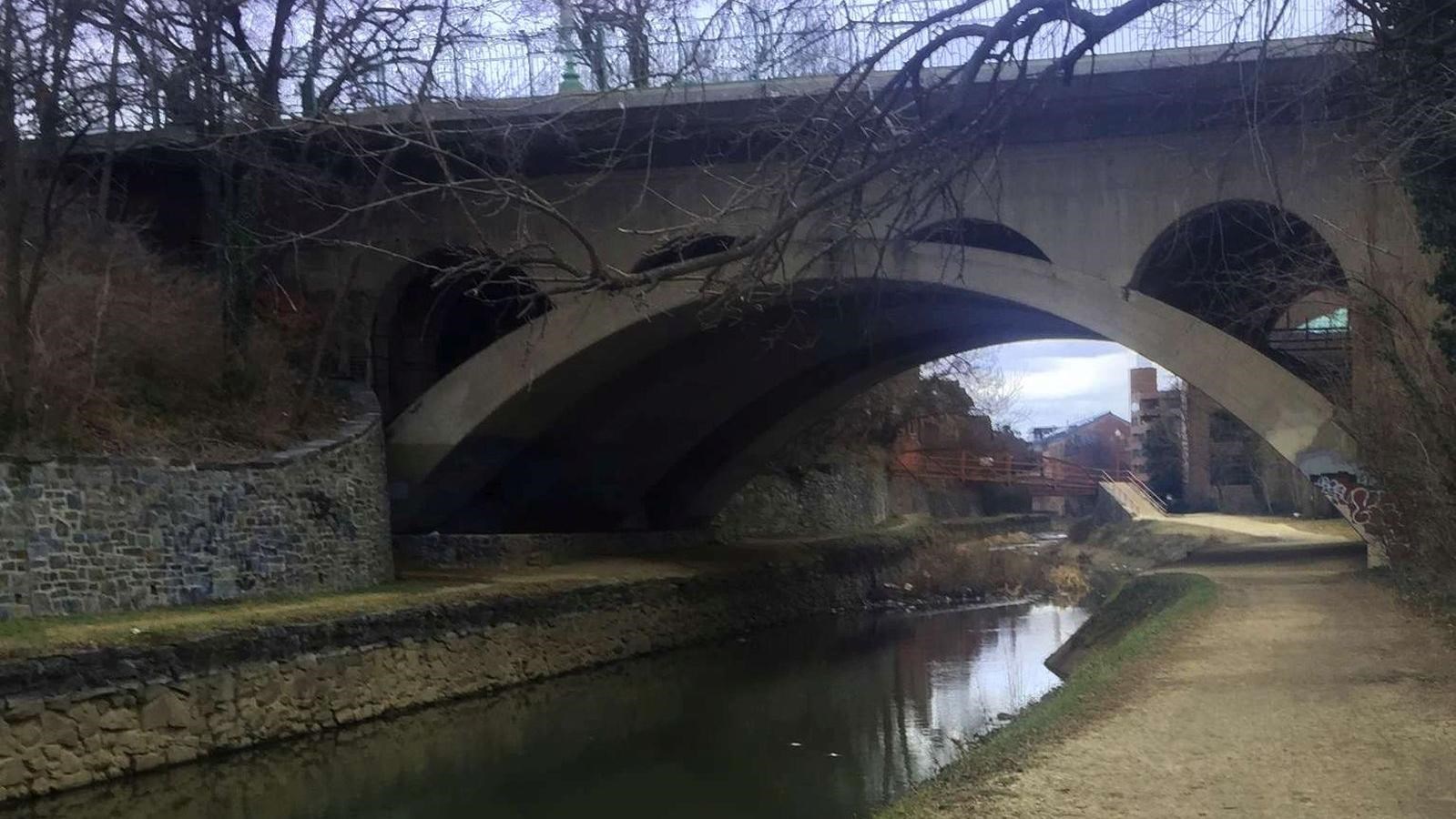 A Concrete bridge arches over a watered canal. A path of loose stone runs parallel to the canal.